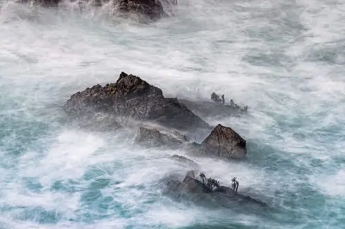 Long exposure photography of rugged sea rocks with small palm trees, surrounded by misty, flowing turquoise and white ocean water.