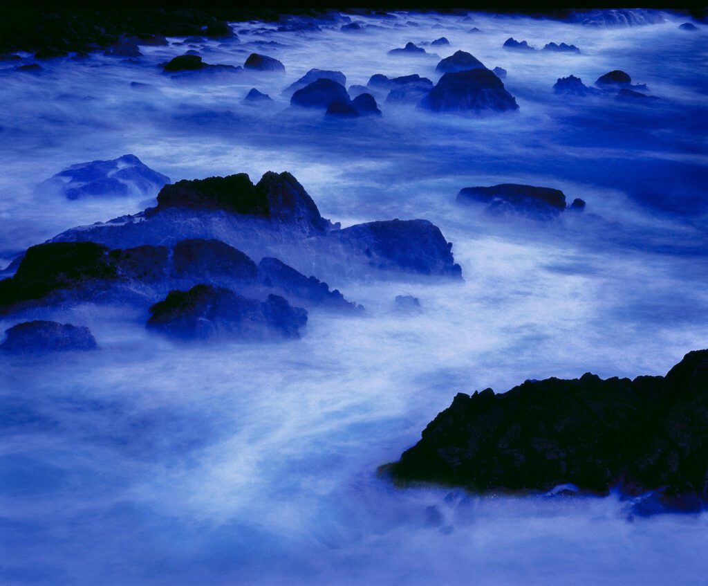 Long exposure captures a dramatic blue ocean scene with dark rocks emerging from misty, ethereal waves, creating a serene and mysterious coastal landscape.