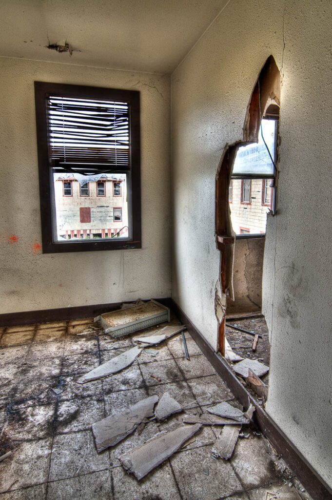 Interior of an abandoned room with cracked walls, broken window blinds, and debris on the floor.