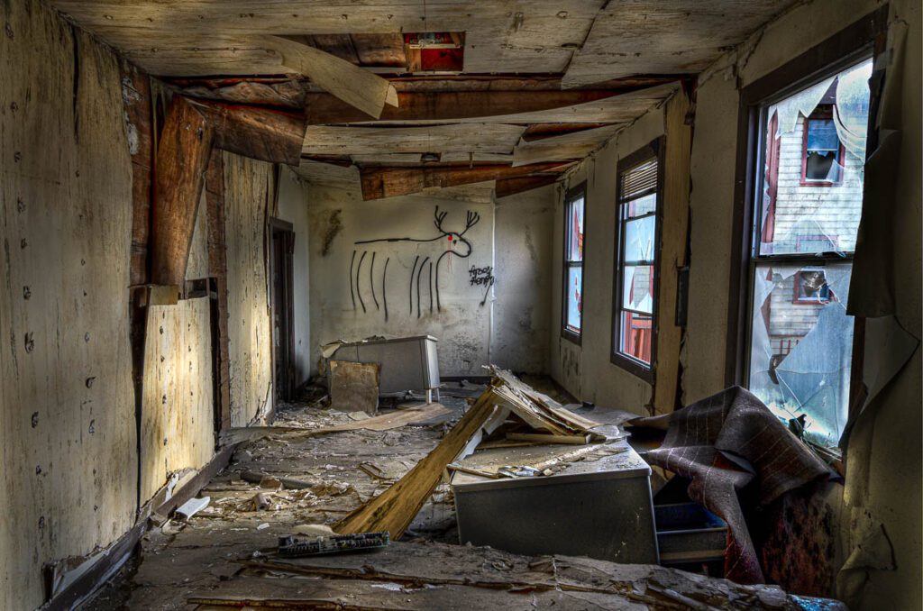 An abandoned room in a modern ruin, featuring peeling paint, shattered windows, debris-strewn floor, and deer graffiti.