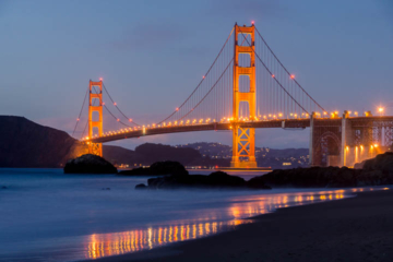 The Golden Gate Bridge as seen from Baker Beach at dusk with lights glowing in the dark blue sky.