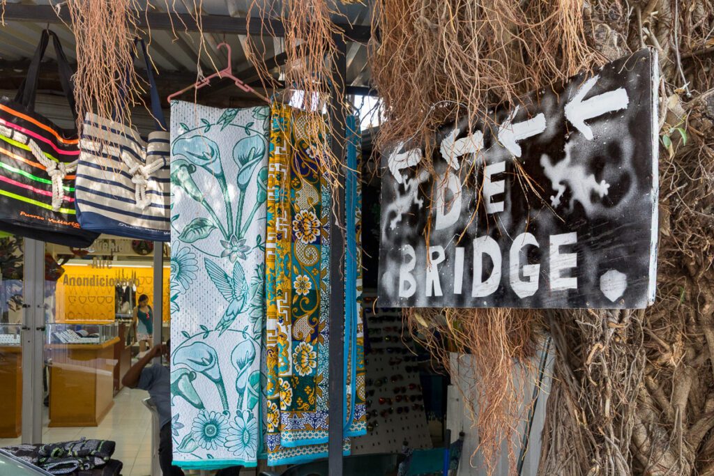 Colorful sarongs, tote bags, and a weathered "BRIDGE" sign with arrows and gecko stencils at an outdoor market stall.