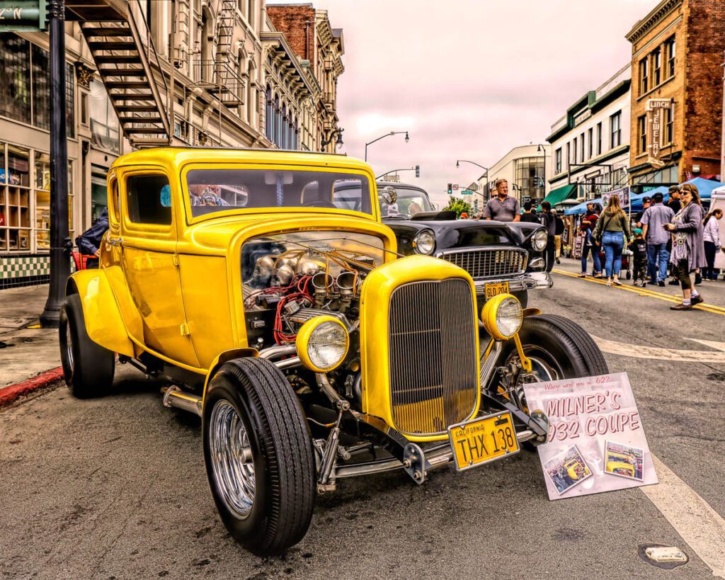 Bright yellow vintage hot rod car displayed at a car show on a street lined with historic buildings and spectators.