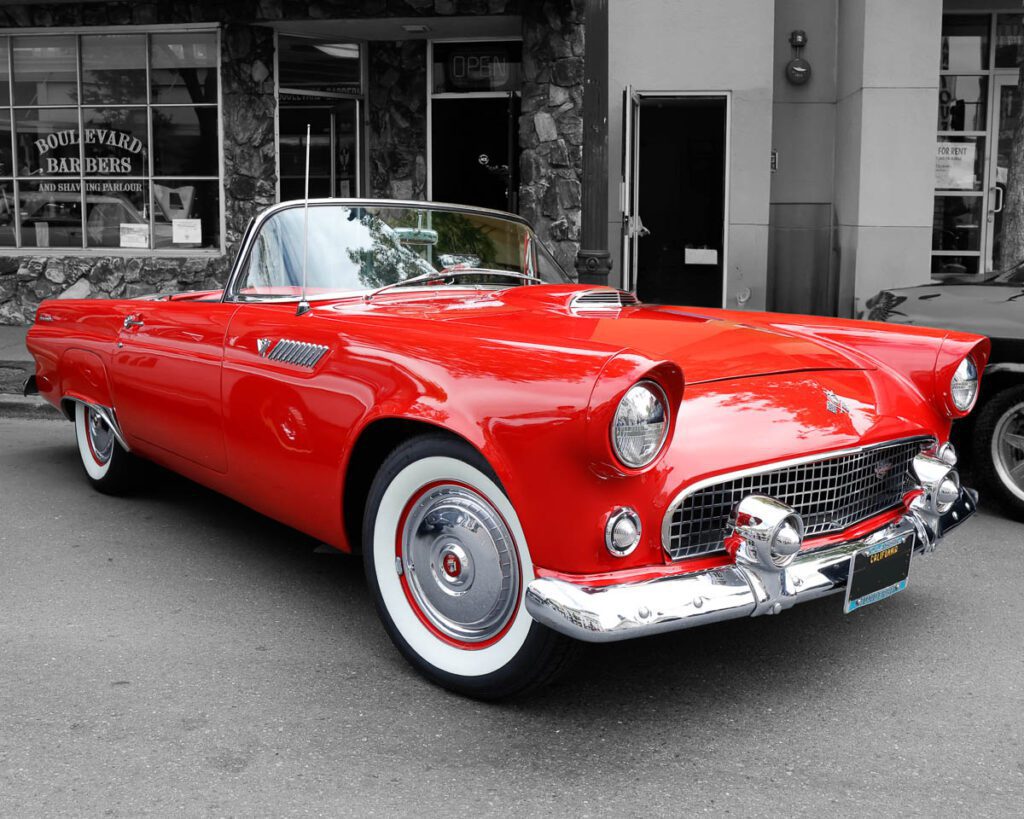 Red vintage Thunderbird parked on a street, featuring whitewall tires and chrome details, with a barbershop in the background.