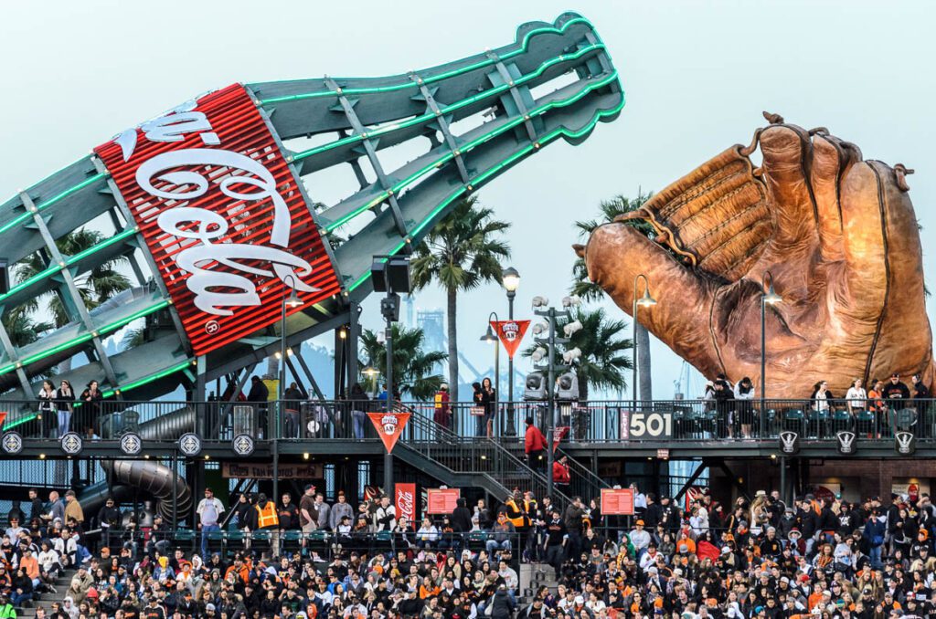 Crowd at San Francisco ballpark with giant Coke bottle and baseball glove sculpture at Oracle Park.