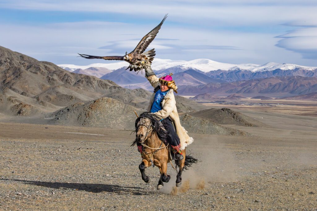 A Mongolian eagle hunter riding a horse with a golden eagle perched on his arm, set against a backdrop of rugged mountains.