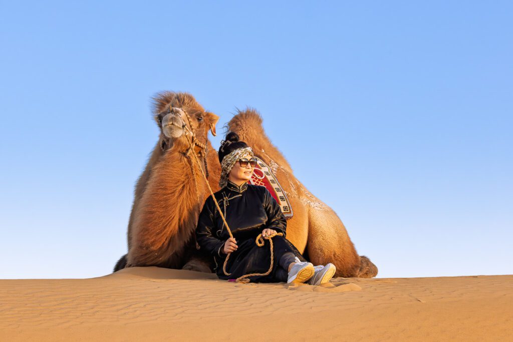Person wearing sunglasses sitting with a camel on a sand dune under a clear blue sky.