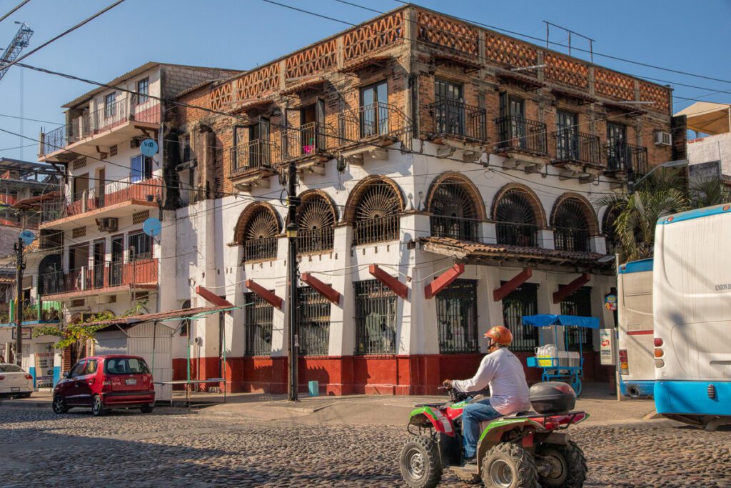 Man on ATV driving on cobblestone street past white and red brick building with balconies and arched windows under blue sky