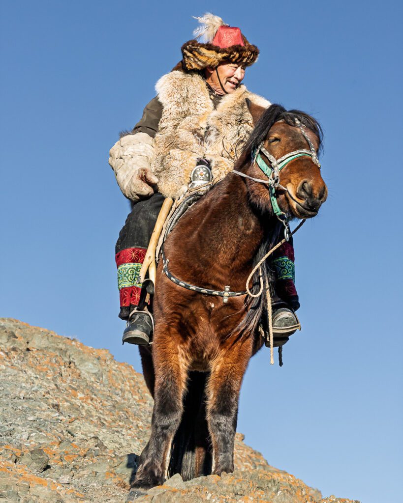 Smiling man in traditional Mongolian winter clothing, including a fur-trimmed hat and coat, rides a brown horse uphill.