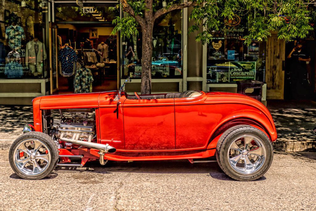 Vintage red hot rod parked in front of storefronts under a tree.